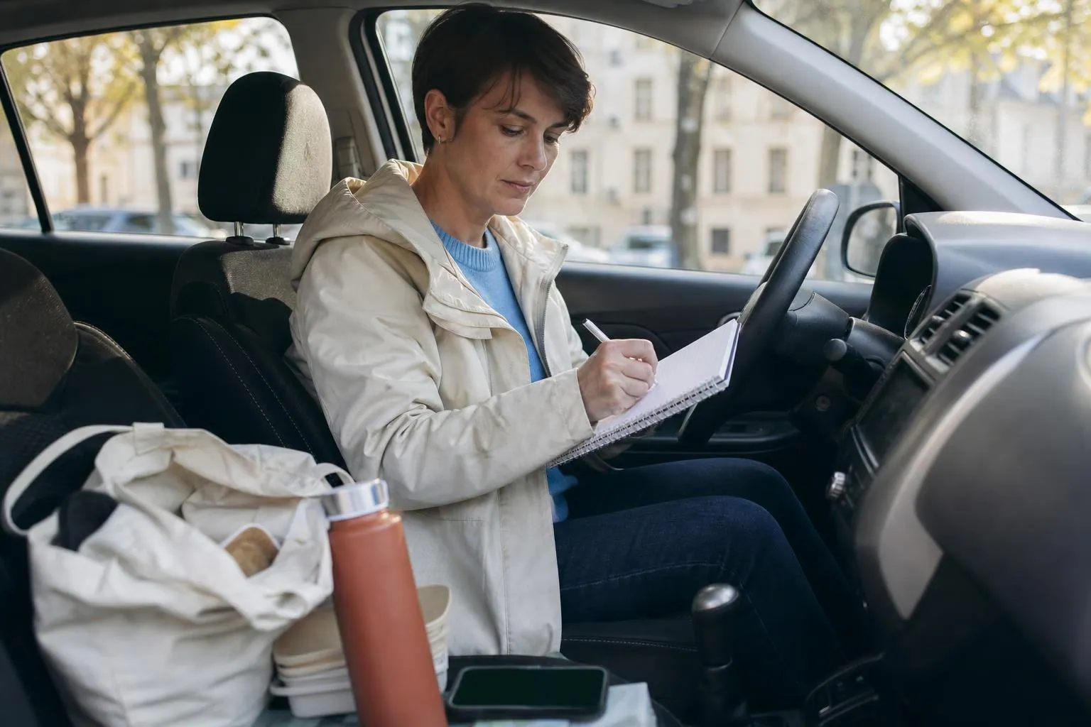 Doula postnatale d’environ quarante ans assise au volant de sa voiture garée devant un immeuble, carnet de notes posé sur le volant et téléphone retourné sur le siège passager